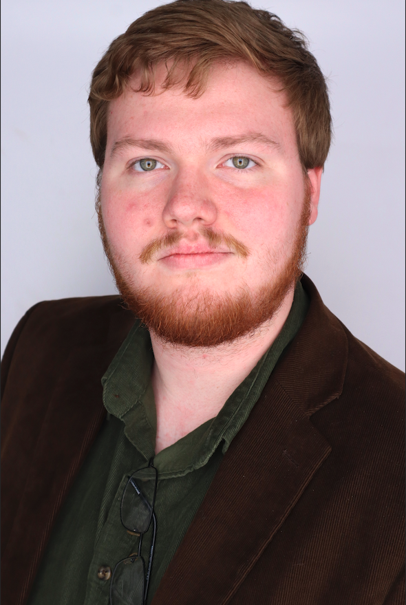 a young man with red hair and glasses, wearing a brown blazer, looks at the camera