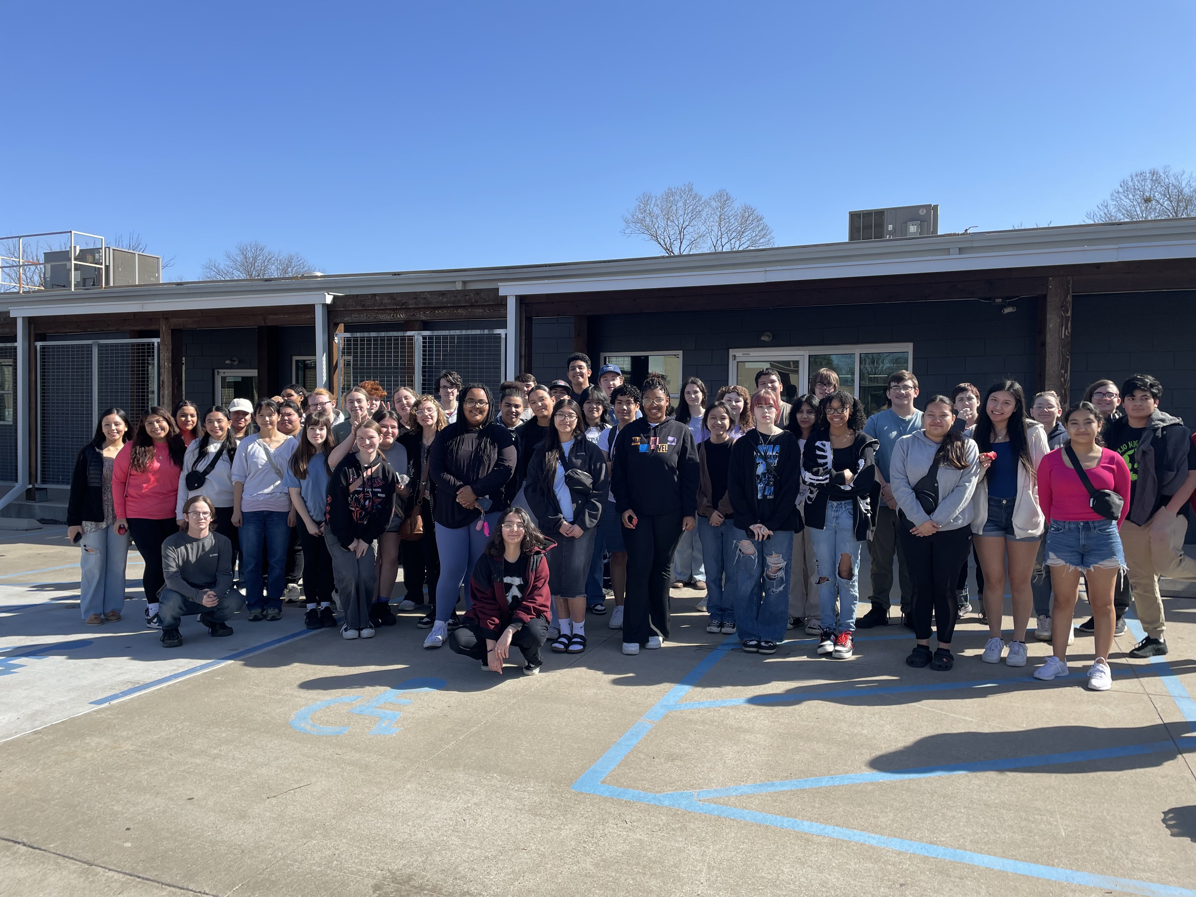 A large group of students stands together outside a building on a sunny day, smiling for a group photo.