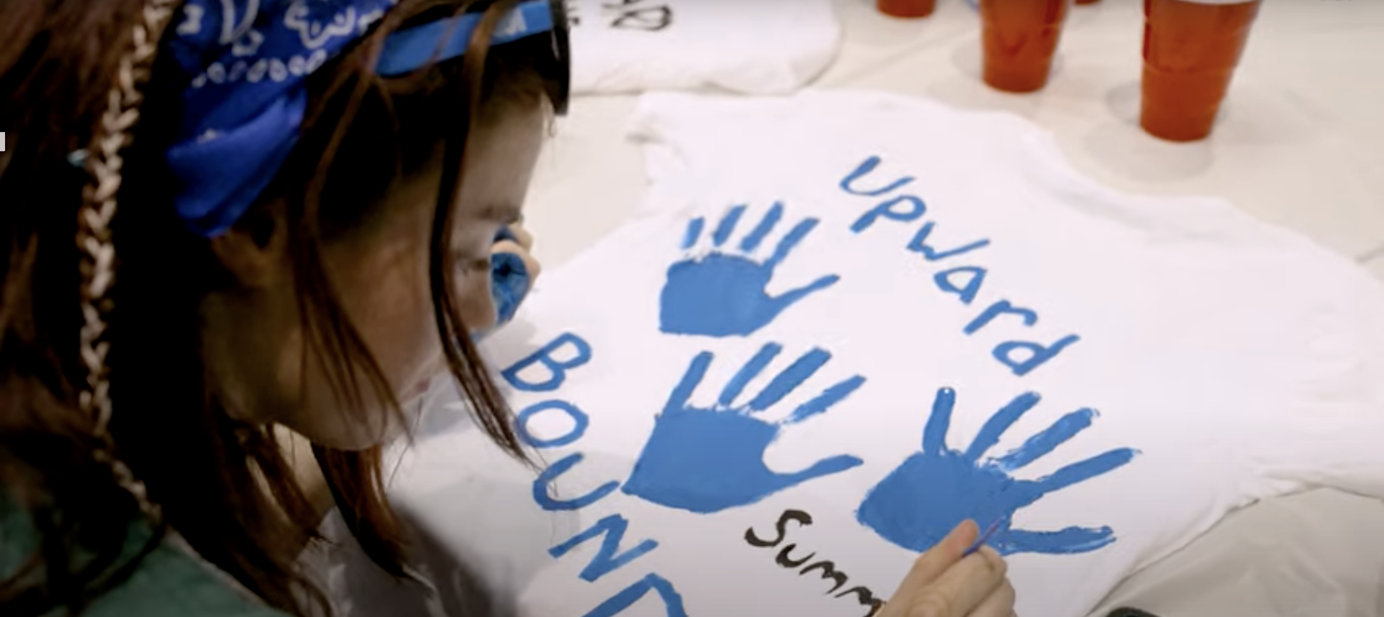A student wearing a blue bandana paints a white T-shirt with blue handprints and the words “Upward Bound Summer.” Red plastic cups and other painted shirts are on the table in the background.