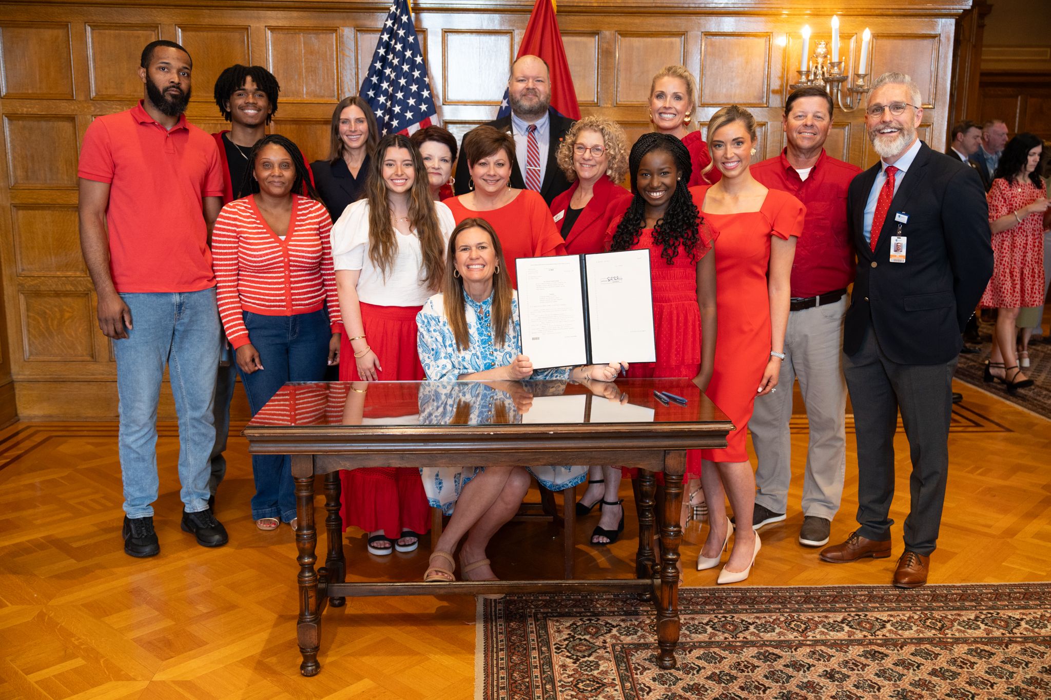 A group of people dressed in red are gathered as Governor Sarah Huckabee Sanders signs a law.
