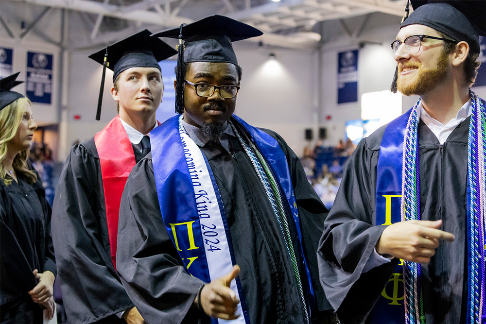 Kendrix Holcomb (center) points to the camera during his graduation in May 2024 in the Stubblefield Center at the University of Arkansas - Fort Smith. He is adorned with his fraternity stoll and Homecoming King 2024 stoll, as well as with multiple cords.