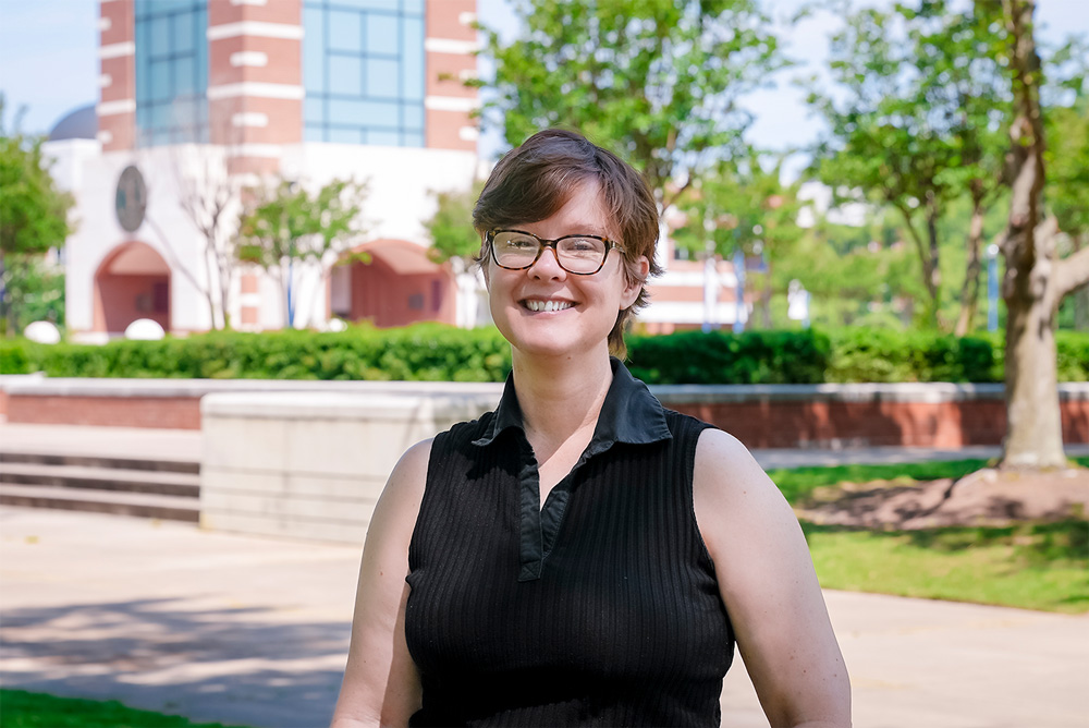 Roxy Wylie smiles while standing on the edge of the campus green at UAFS with the bell tower behind her