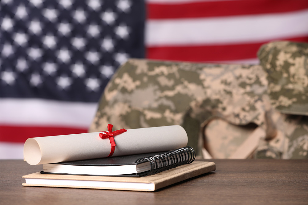 Image of an American Flag (background) with a stack of two notbooks and a diploma sitting on a desk in front of a military uniform draped over a chair